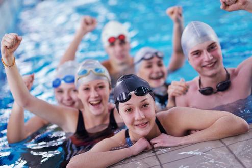 Sechs Jugendliche in Badekappen und Schwimmbrillen posieren fröhlich im Wasser, einige mit erhobenen Armen.